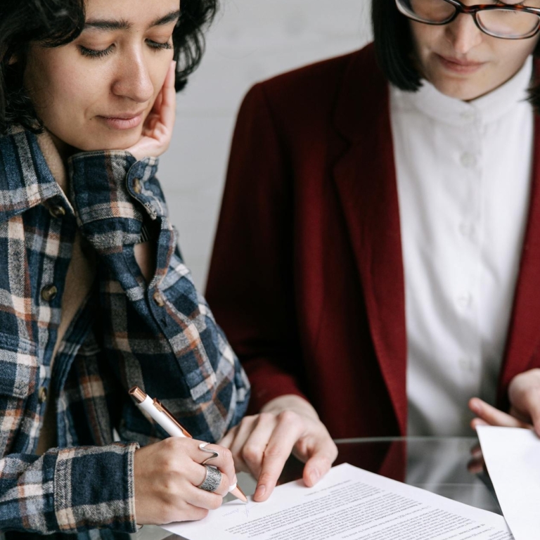 Two women reviewing and signing documents together at a glass table indoors.